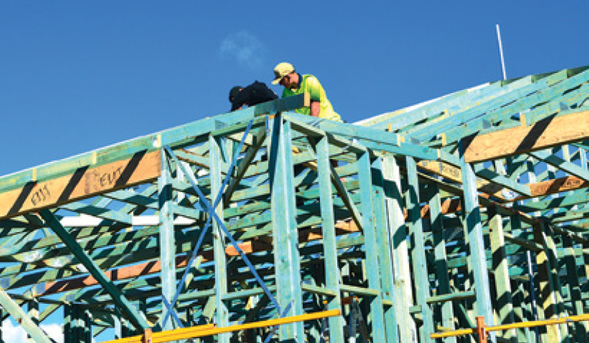 A construction worker on his knees on the roof of a timber-framed house. The worker is wearing a bright yellow high-visibility vest, a hat, and carries a work bag.