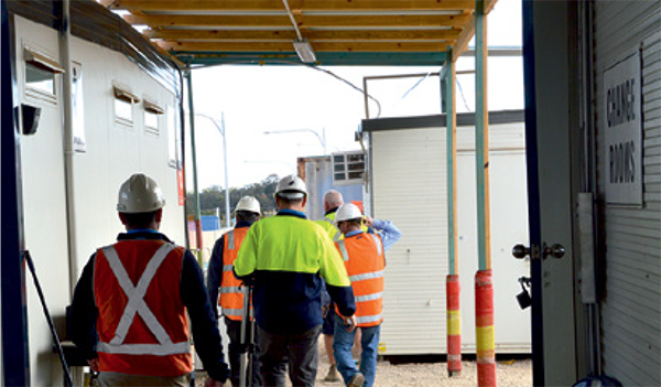 Group of workers wearing different high-visibility vests and white hard hats at a construction site walkway access point, near what appears to be an office and rest area.