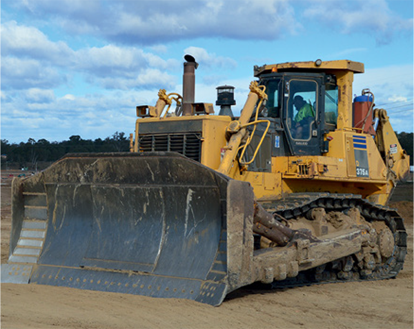 A yellow bulldozer is at a construction site with a worker driving it, wearing a high-visibility vest. Trees are visible in the background.