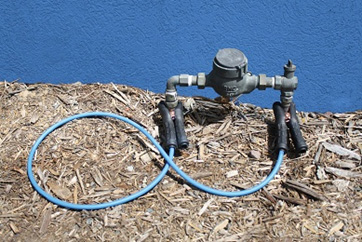 A water meter with a blue bridging conductor clamped to it. The background features a blue wall, and the ground is covered with wood chips