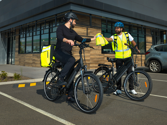 One food delivery rider wearing a high vis vest, passing a vest to another food delivery rider. 