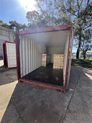 An open red shipping container with white interior and doors wide open, another open container visible in the background, surrounded by trees and grass.