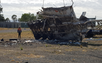 A cryogenic tank container and skel trailer after a rupture, with the tank shattered into multiple pieces and lying on its side, resembling an explosion. A person wearing a helmet and high-visibility vest is inspecting the wreckage.