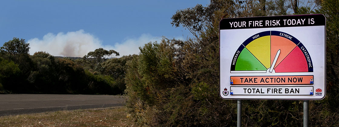 A fire risk road sign on the side of a road, with smoke in the distance. The risk rating is pointing to 'extreme'. 