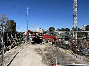 South-facing view of the rear of a red concrete pumping truck at a construction site, tipped over on its side behind a wire fence