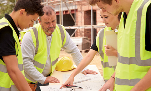 Group of construction workers wearing high-visibility vests gathered around a table at a building site. A hard hat is on the table, and one worker is pointing to a section of the building plans as they review them together.