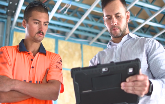 Two workers looking towards an iPad on a construction site. The worker on the left is wearing high vis, the worker on the right is wearing a corporate shirt.