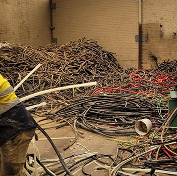 Inside a warehouse with yellow brick walls, piles of black and red cables are scattered on the ground. On the left, the back of a worker wearing a high-visibility vest is visible.