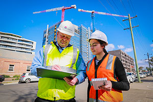 Two SafeWork inspectors wearing PPE and holding clipboards, on a construction site.