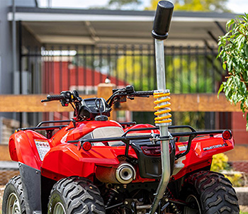 A red quad bike is parked showing it's back view. It has a Flexi, a metal bar with a black handle at the end. The middle section features a yellow spring suspension. The background included a carport and a fence.