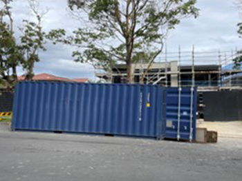 A blue shipping container positioned in front of a construction site near a road, with houses and trees on either side.