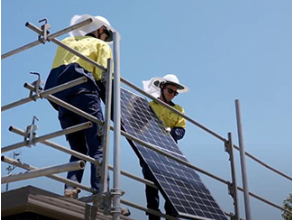 Two workers in PPE, installing a rooftop solar panel. There is a temporary guard rail on the edge of the roof.