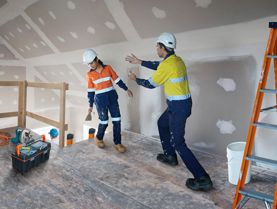 Two workers wearing hard hats and high vis, pictured near a void. One worker appears to be stumbling backwards, with the other worker attempting to reach them.