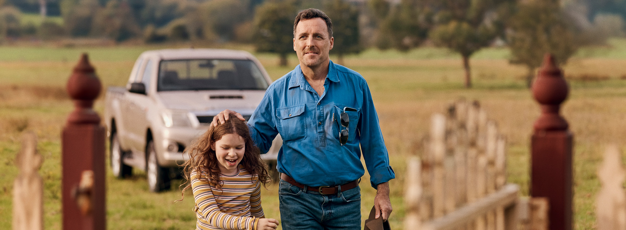 Farmer wearing a blue collared shirt tenderly touching his daughter's head as they walk through their farm