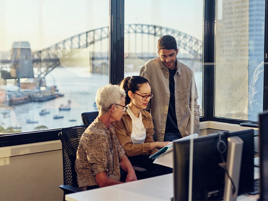 A group of young workers looking at a computer screen together. The Harbour Bridge is in the background.
