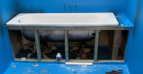 A bathtub is set within a wooden framework, with blue sealer painted around it. Various tools are scattered on the ground.