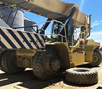 A yellow 60‑tonne reach stacker is shown on a dirt surface with its front tyre removed and resting on the ground beside it.