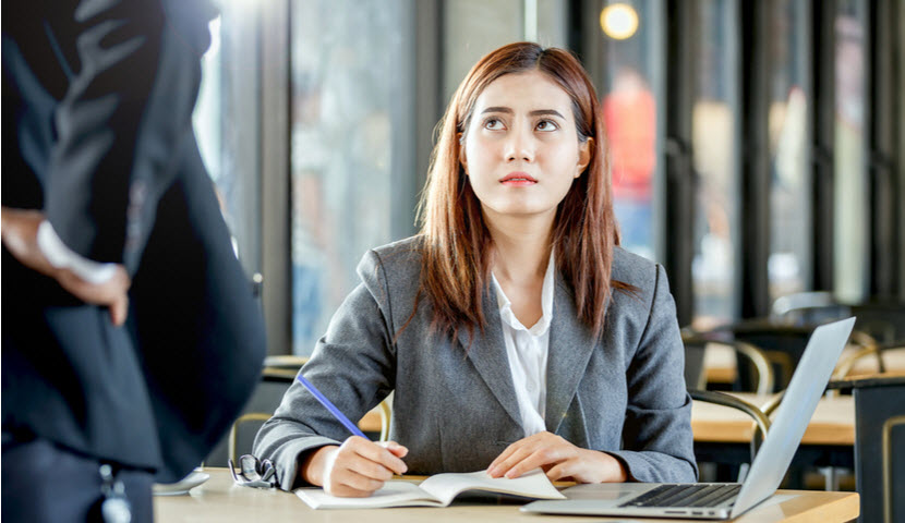 Upsetting face expression of young female employee sitting and writing note on table with laptop in front of angry manager