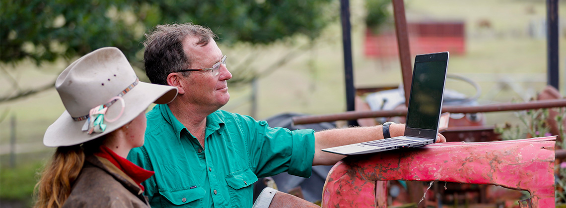 Two people on a farm, with one resting a laptop on a piece of machinery