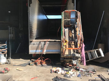 The rear if a truck with its back doors open. An elevated working platform is positioned to the right of the truck.