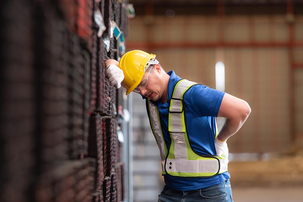 Male worker wearing a safety vest and helmet standing on a steels pallet due to back pain from working in a factory lifting heavy things.