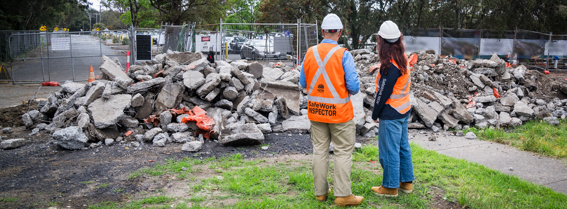 Two SafeWork inspectors wearing orange high-vis vests and white hard hats inpsecting rubble on a work site.