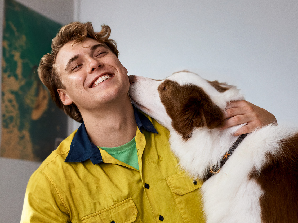 Apprentice wearing yellow shirt smiling as his brown and white dog sniffs his neck